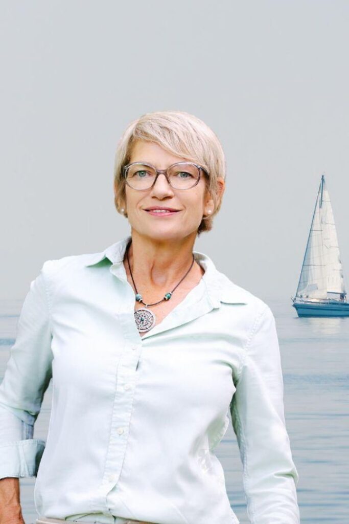 Coach Caroline Schmitt steht lächelnd und gelassen vor einem maritimen Hintergrund mit einem Segelboot auf dem Wasser.